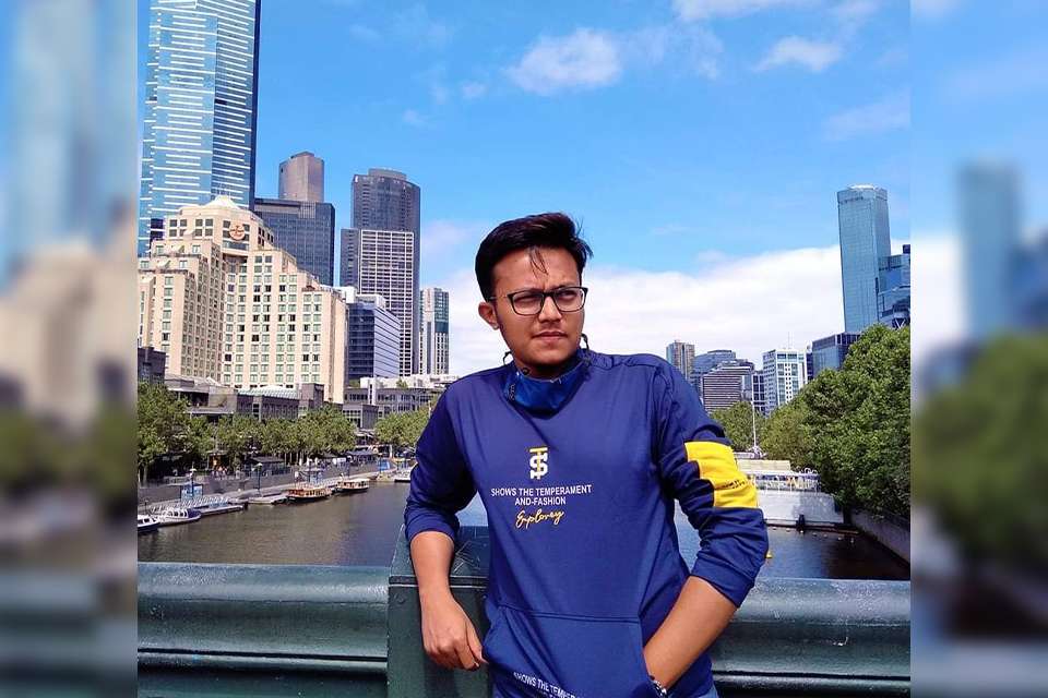 Jaber Alam stands on a bridge over a river, with the city of Melbourne as a backdrop behind him as he looks off into the distance introspectively.