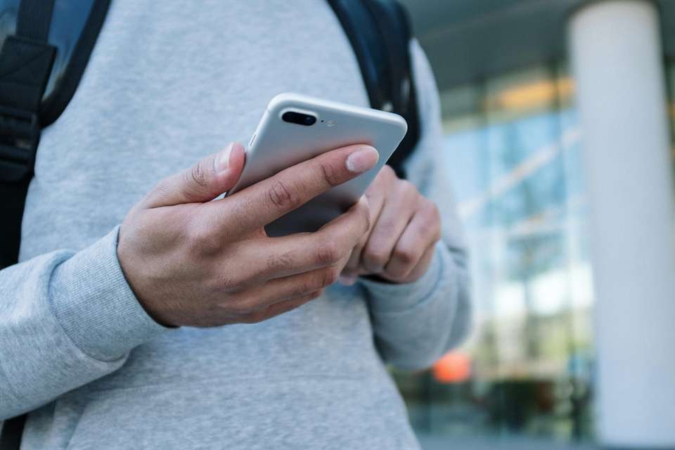 A close up of someone holding a mobile phone in their hand. The phone is silver and you can see that the person is wearing a grey sweater and a backpack.