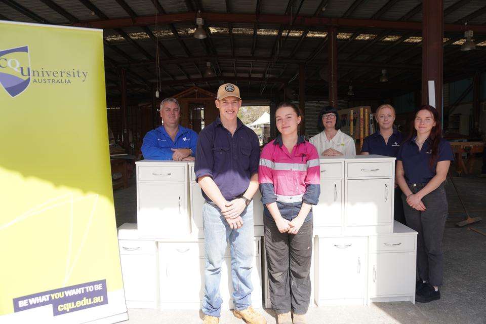 A group of 6 people comprising of students, a teacher and stakeholders stand in a trades workship with 11 bedside tables and a CQUniversity pull-up banner.