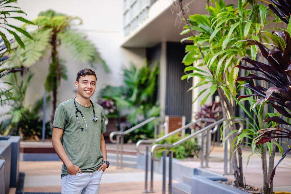 Medical student Jay Warcon stands amidst tropical campus gardens, as he smiles at the camera with a stethoscope around his neck