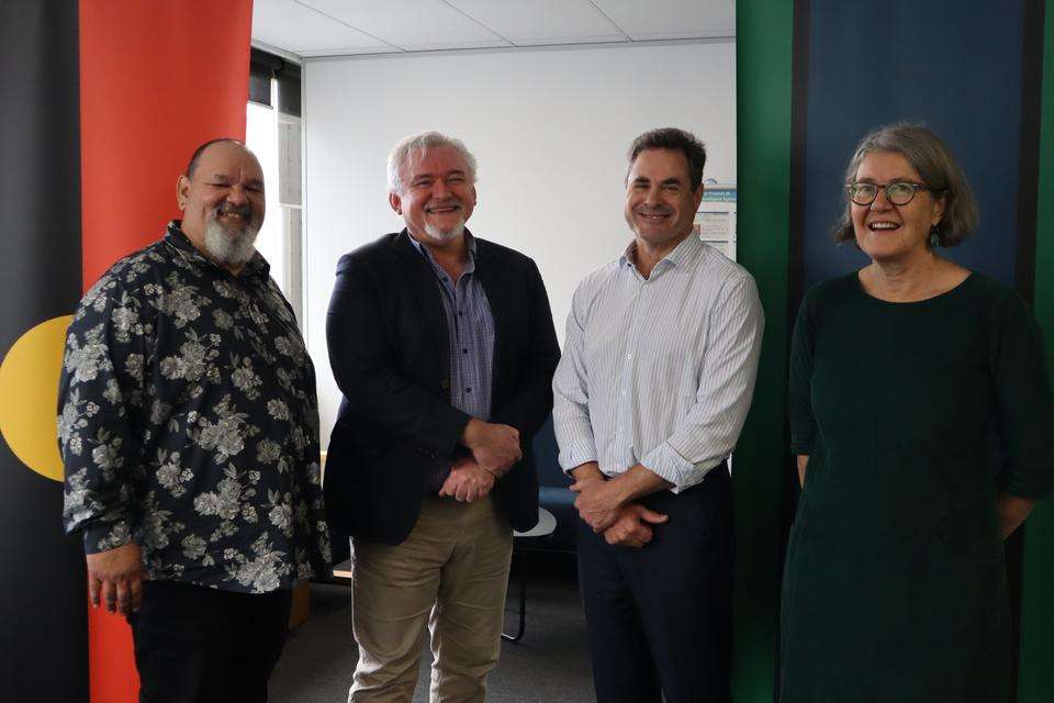 Researchers from Jawun Research centre celebrating establishment of the Jingay (pronounced Jin-guy) research cluster. People in image include from L-R Professor Adrian Miller, Greg Pratt (head of Jingay),Professor Grant Stanley and Dr Janya Mccallman. The group is standing in front of two corporate style banners, one featuring the Aboriginal flag and the other featuring the Torres Strait Islander flag.