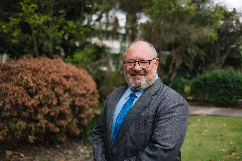 A man wearing a suit, blue tie and glasses stands outdoors.