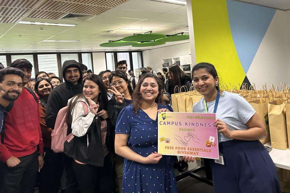 A group of students and staff stand in front of a table holding dozens of brown paper bags. Staff hold sign reading Campus Kindness Project.