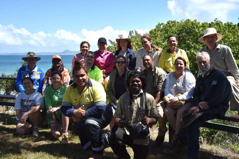 A group of conservation volunteers sit together smiling for a photo; they sit against a backdrop of greenery and ocean views.