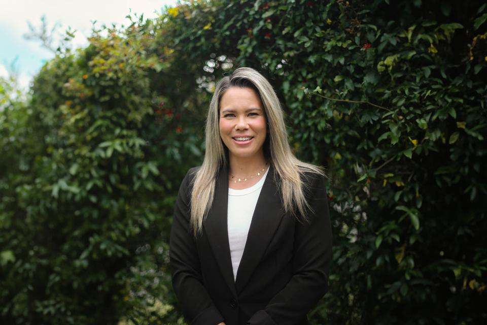 Lauren Turner stands in front of a tall green hedge. She has long blonde hair and wears a white shirt and black jacket.