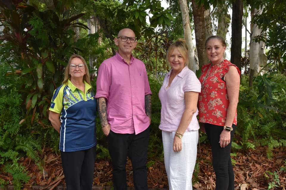 Four teachers stand outside against a backdrop of native greenery; they are standing proudly and smiling, and are dressed colourfully.