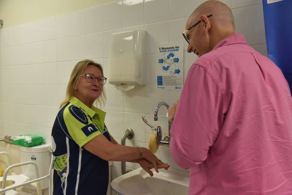 Two teachers stand in a simulated hospital ward at a basin as they demonstrate hand-washing techniques. They are looking at each other engaged in happy conversation.