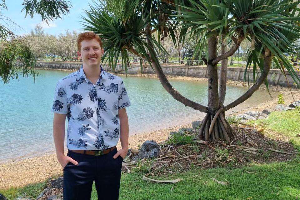 Logan Crosby stands by a beach location with a palm tree and sea wall in the background.jpg