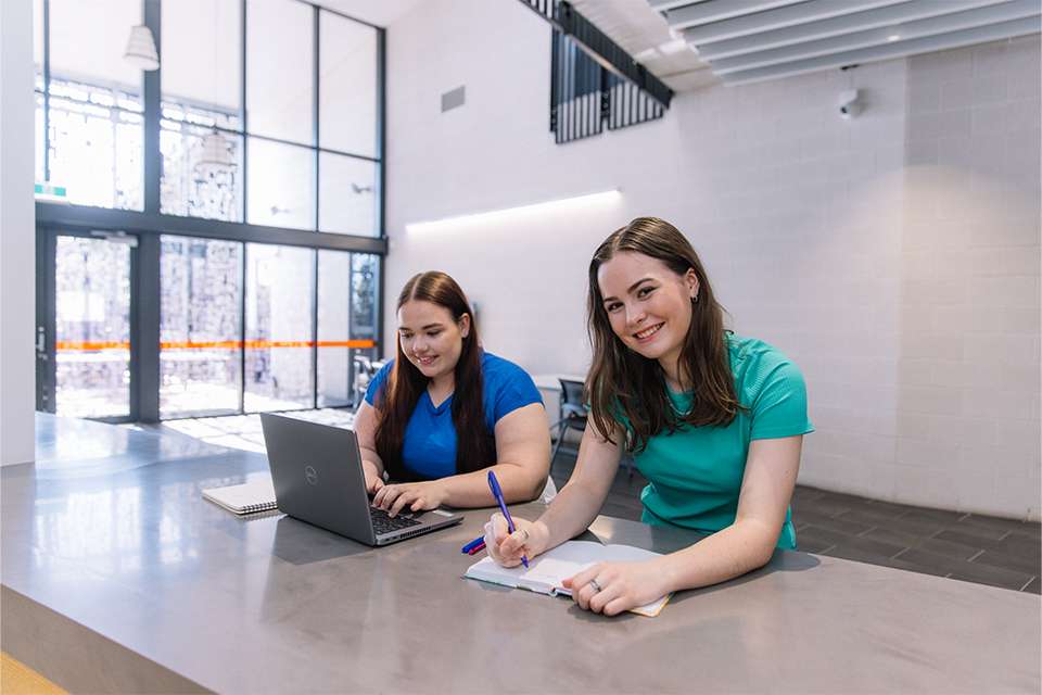 Two students studying on campus, one working on laptop and another writing in notebook and smiling at camera