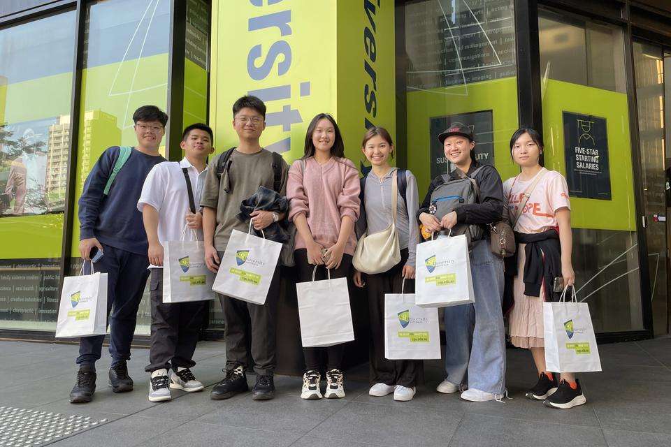 A group of young men and women of Asian appearance standing in front of a CQUniversity sign and holding tote bags