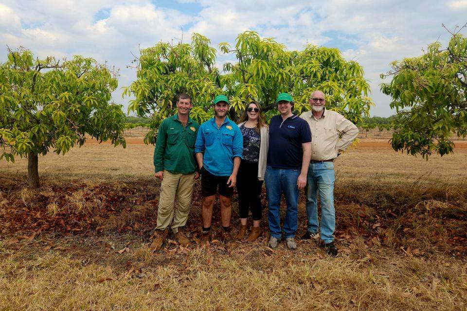 Five people all looking directly at the camera and smiling while standing in front of a small mango tree on a farm with dry brown grass