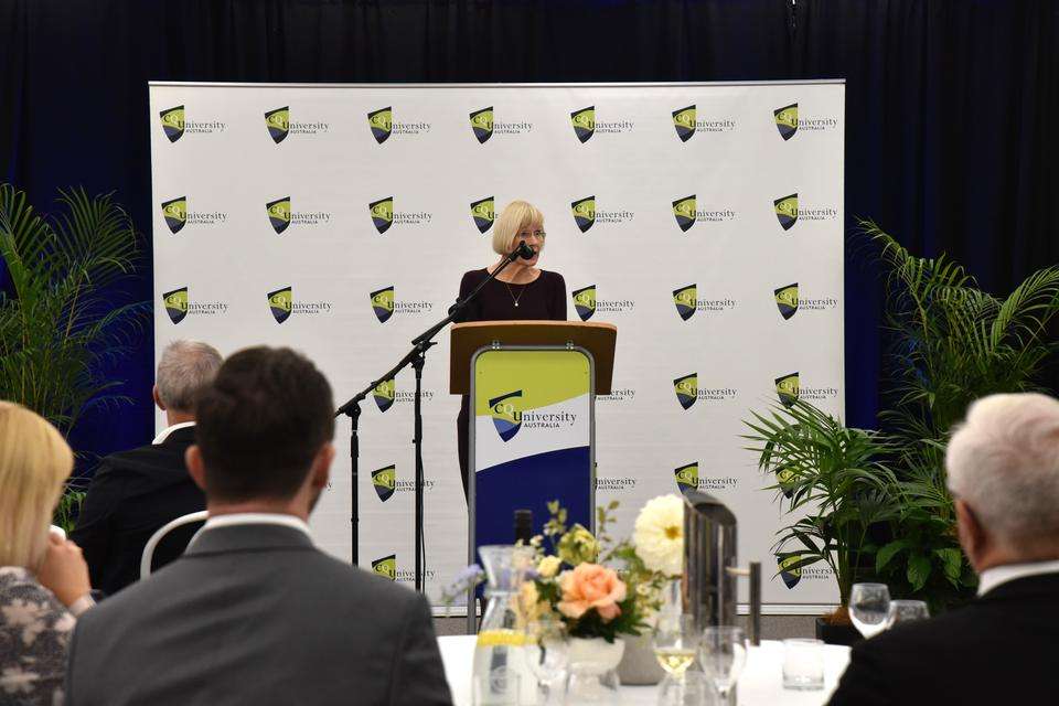 UQ Vice-Chancellor and President Deborah Terry AC stands to give a speech at a CQU podium with a spotted CQU media wall in the background, stakeholder guests sit listening in the foreground