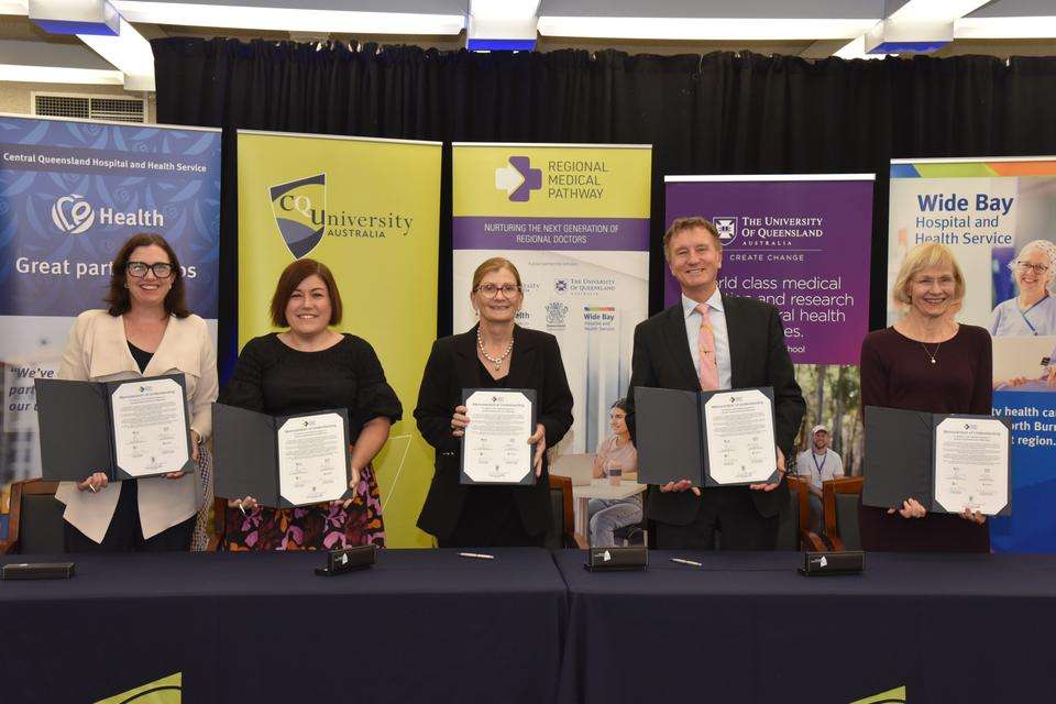 Stakeholders of CQU, UQ, CQHHS stand smiling in front of representative media banners, holding signed Memorandums of Understanding