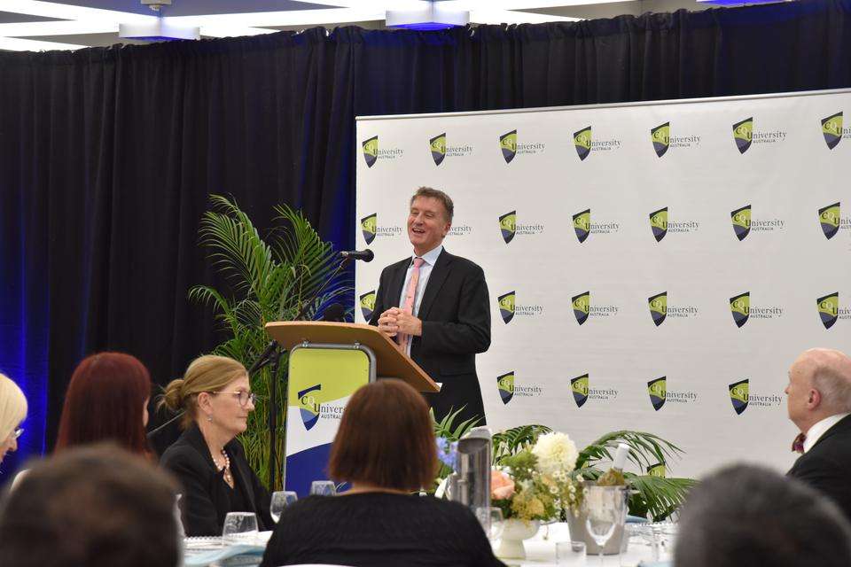 Vice-Chancellor and President Prof Nick Klomp stands to give a speech at a CQU podium with a spotted CQU media wall in the background; stakeholder guests sit in the foreground