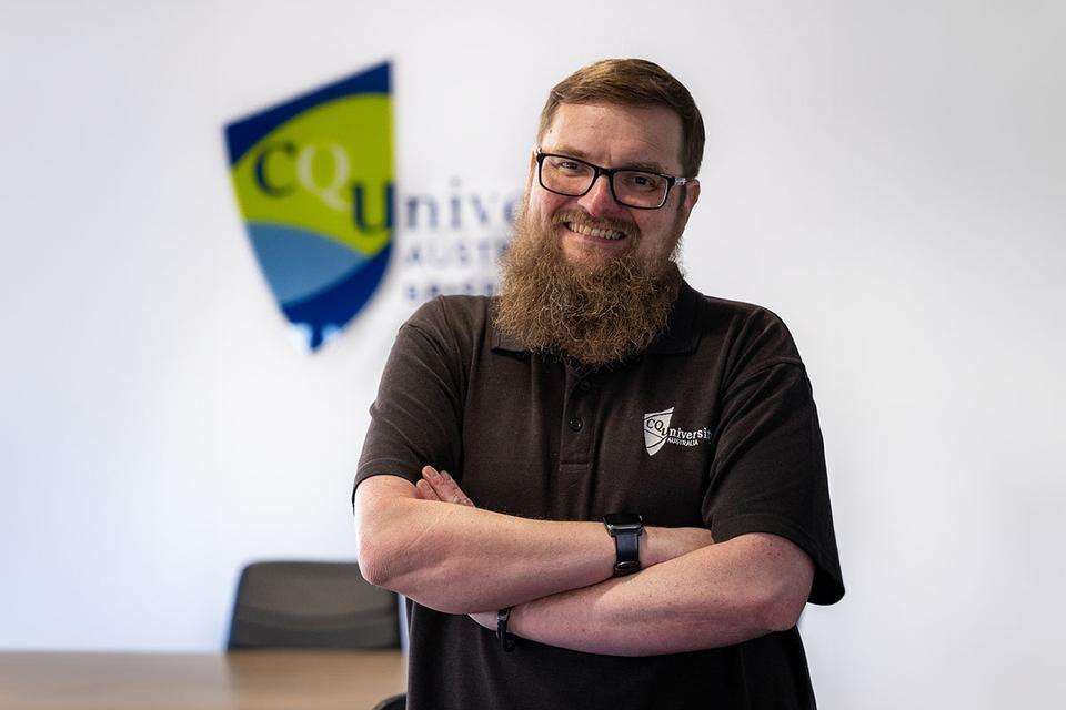 Michael Cowling stands with arms folded in front of a table and CQUniversity logo on the wall.jpg