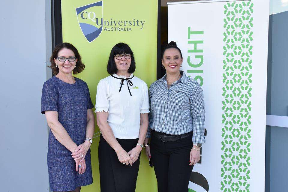 Three women in corporate wear smiling to the camera with CQU and MyneSight banners behind them