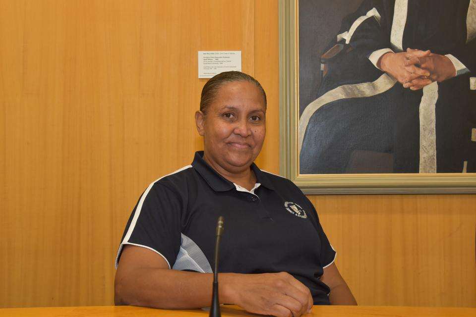 A First Nations woman sits at a speaking desk, smiling at the camera, part of a portrait of a vice-chancellor hangs in the background on a wooden wall