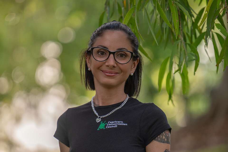 Conservationist and Environmental Scientist Sophie George smiles as she stands outside under a tree. She wears a Capricorn Conservation Council t-shirt and black-framed glasses.