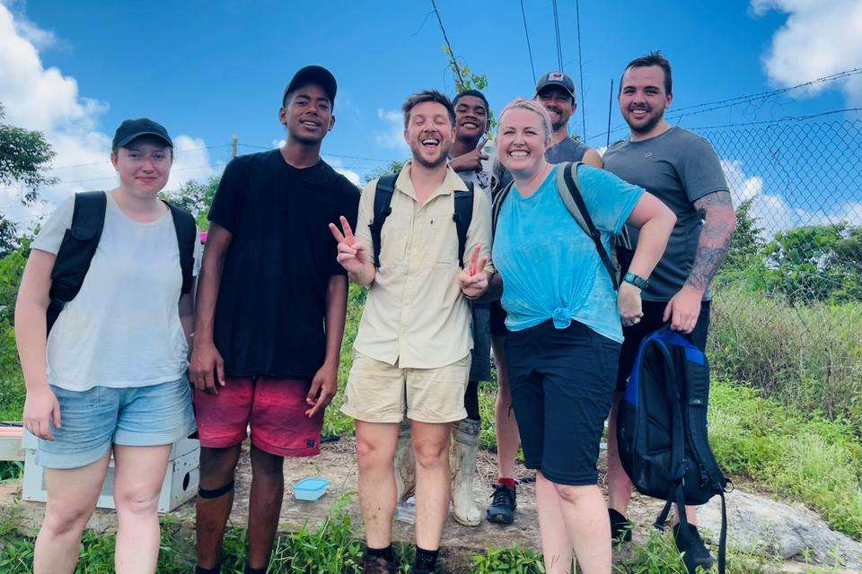 A group of young people stand smiling for a photo outside amongst bushland, they are geared up for a trek with boots and backpacks.