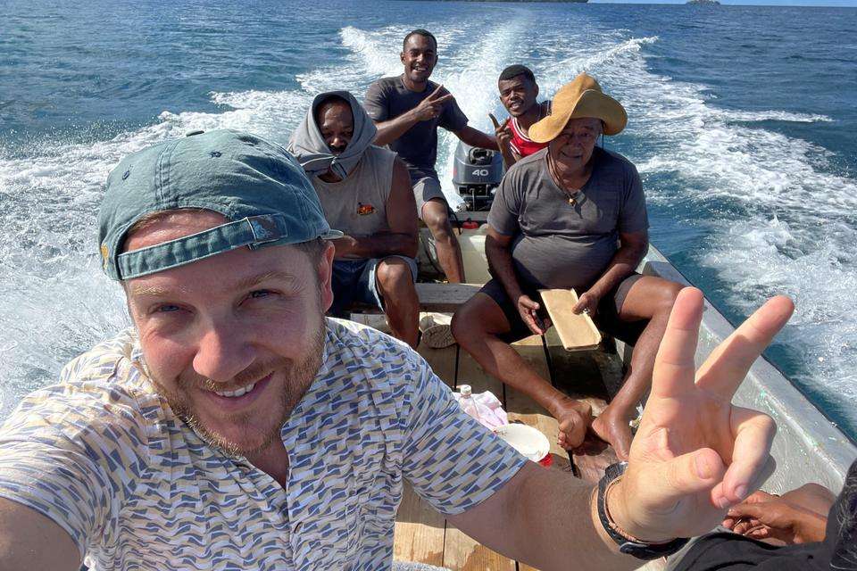 Jake Gries is pictured on a small boat with local Islanders, the expanse of blue sea and mainland behind them. They smile and make the peace symbol with their hands.