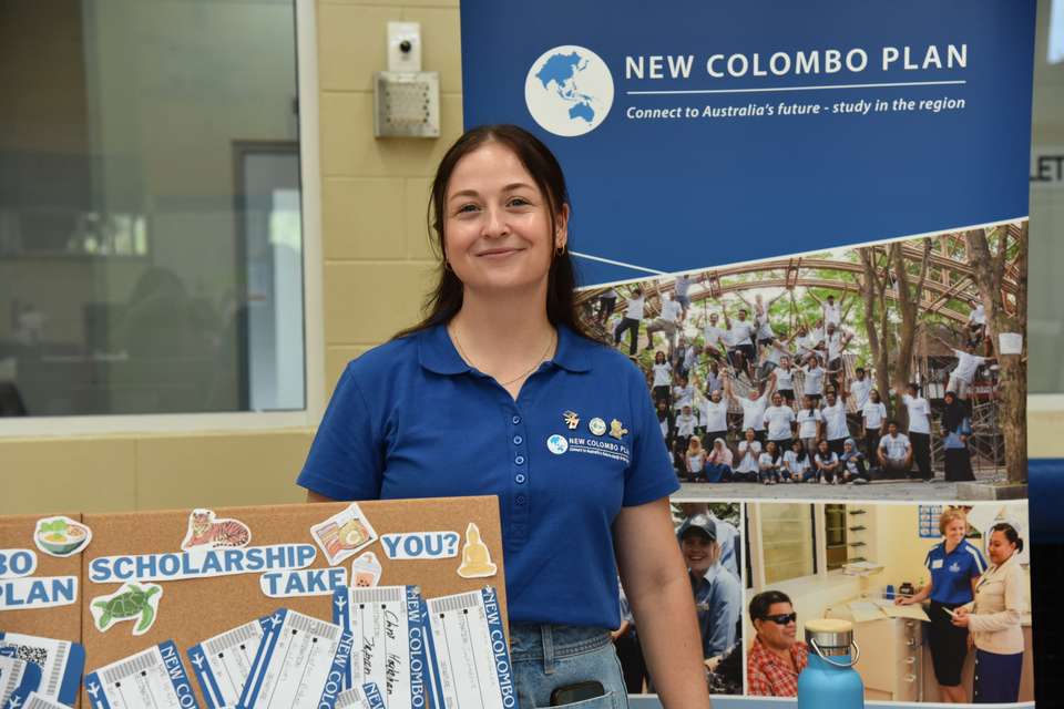 New Colombo Plan Scholar Carlie Sommers stands at an NCP information stall, decorated with a branded pull up banner and pretend boarding passes on a pinboard to advertise learning abroad opportunities.