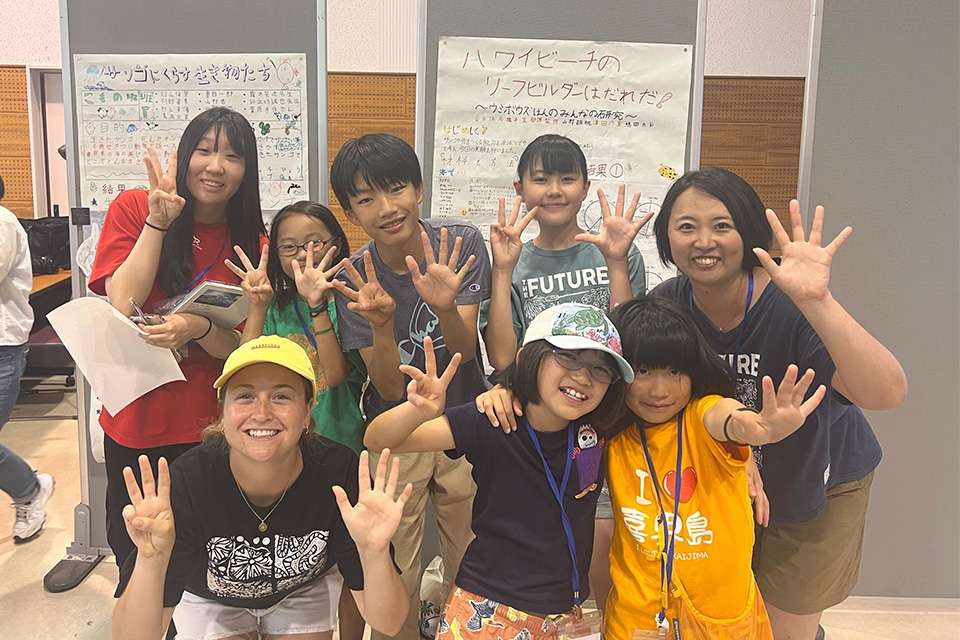 New Colombo Plan Scholarship recipient Carlie Sommers stands posing and smiling with a group of Japanese teachers and primary school students; they each hold up eight fingers to signify something of meaning.