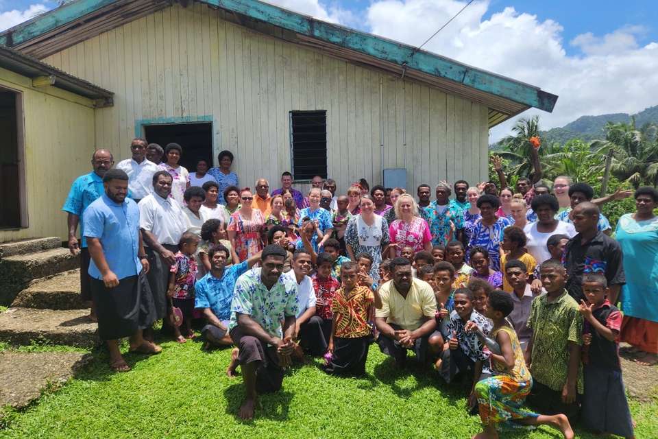 A group of nursing students and Fijian villagers all in colourful clothing stand together outside for a group photo. An old shack and palm trees frames as the backdrop.