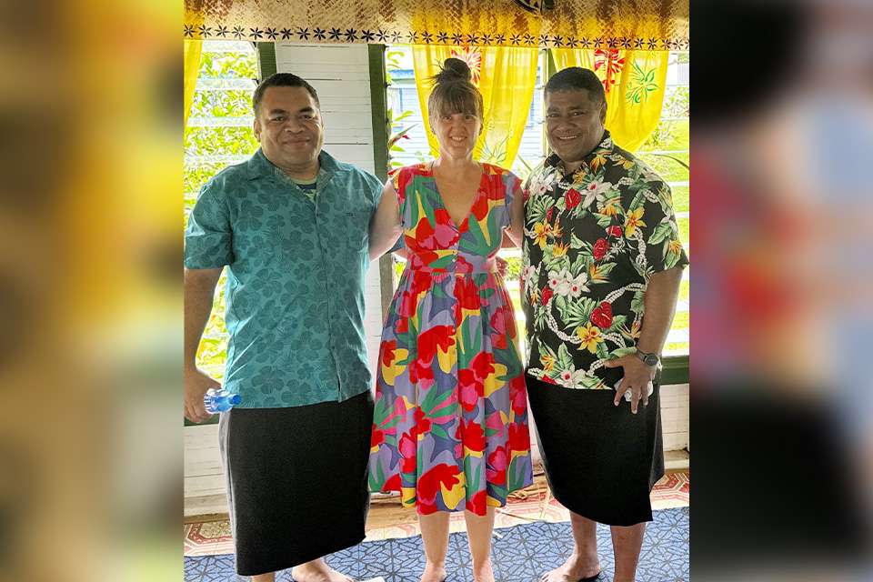 Three people wearing colourful clothing stand together smiling for a photo, in a colourful room.