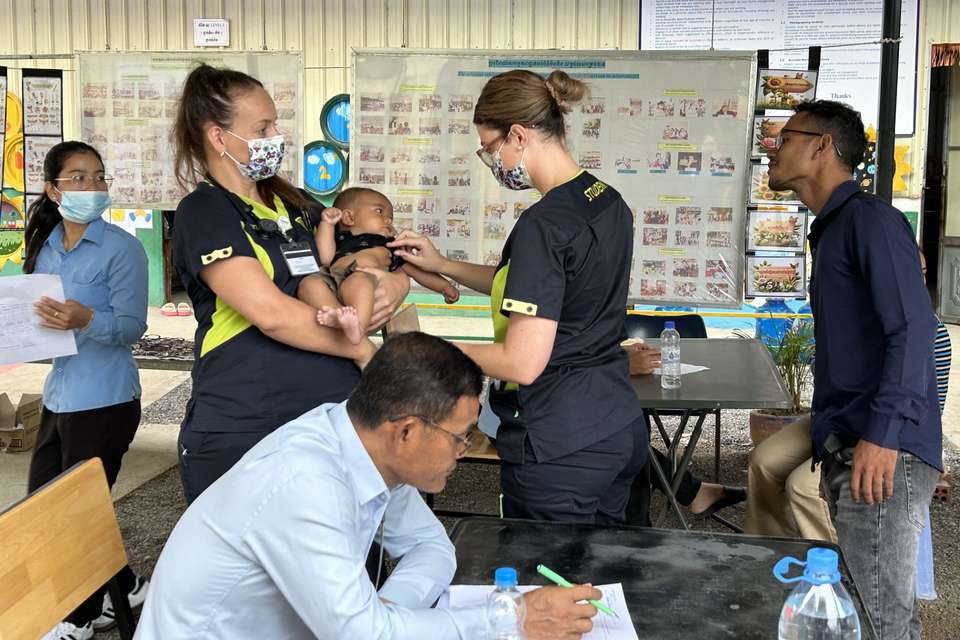 Two nursing students perform a health check on an infant, with other medical professionals working around them in a health facility.