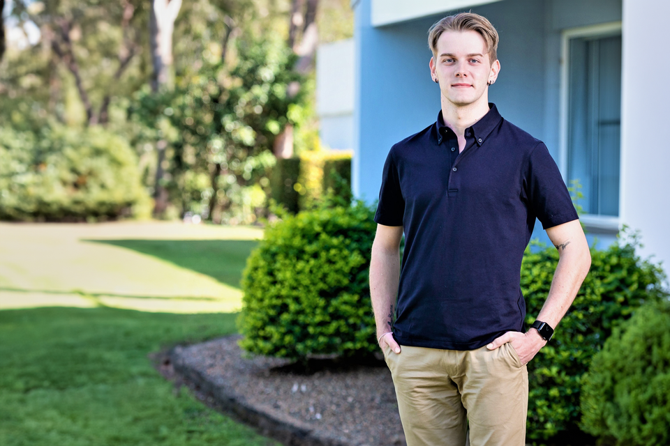 A young man with light coloured hair pictured standing alongside a building and garden on the Bundaberg campus. He is dressed casually in a dark blue collared shirt and beige pants.