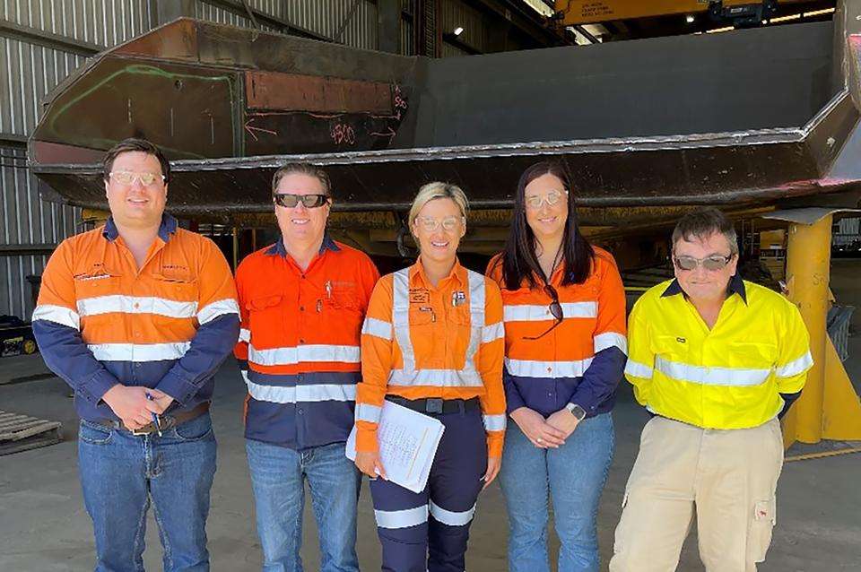 A group of five men and women in hi-visibility clothing and wearing safety glasses stand in a workshed smiling at the camera