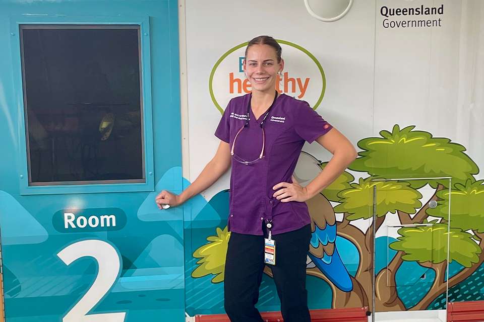 Oral Health Therapist Olivia Arnold stands smiling outside a colourful school dental van where she works.