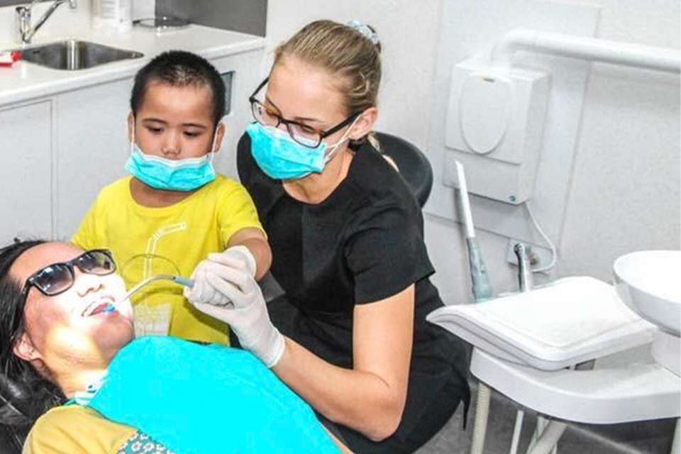 Olivia Arnold in a dental clinic treating a patient; she guides the patient's son with a mouth suction
