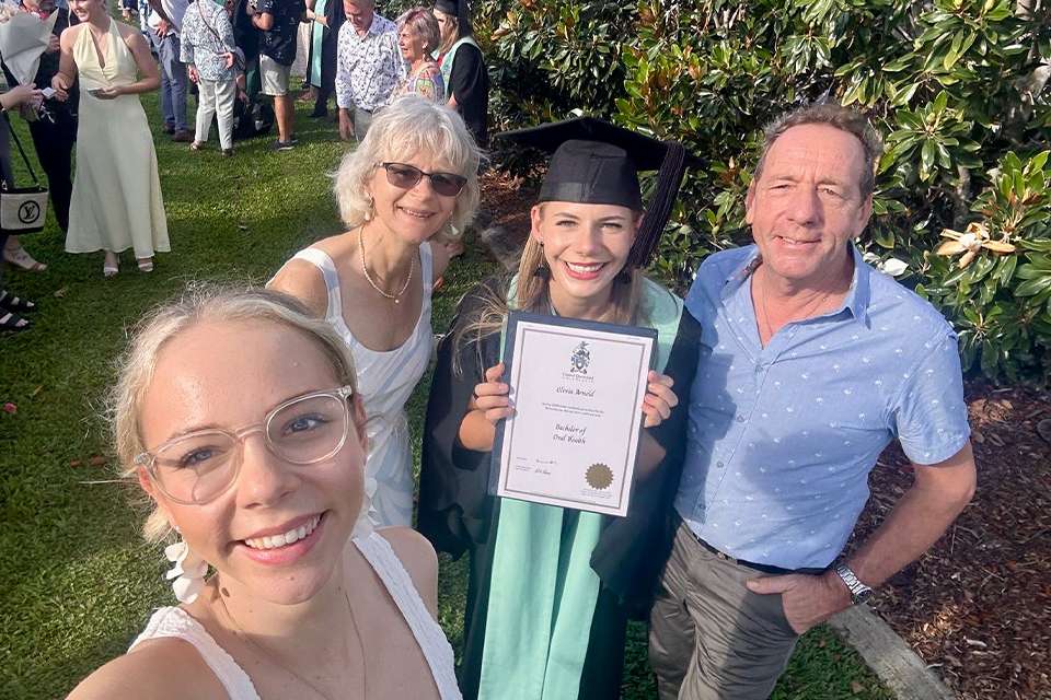 A happy graduate stands outside with her family; she is smiling and holding her testamur