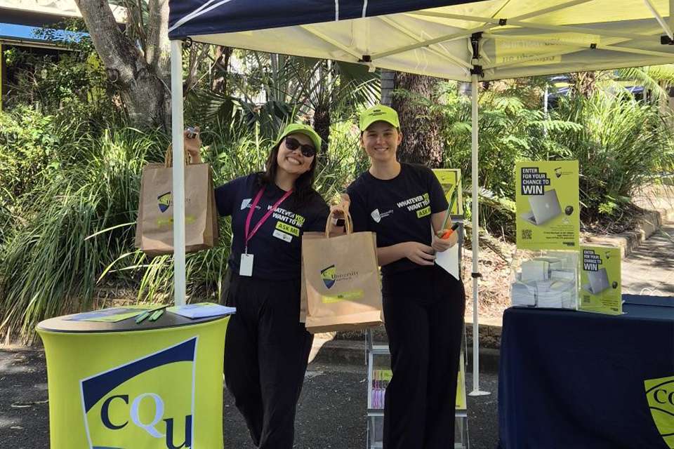 Two CQU staff stand outside on campus, smiling and dressed in CQU-branded clothing and caps at a marketing stall with goodie bags and flyers
