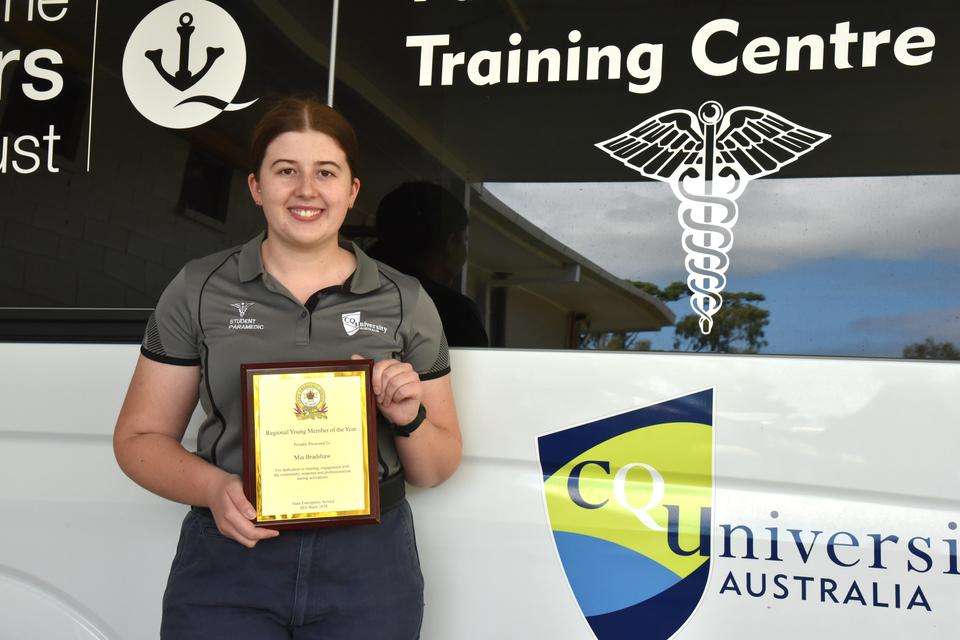 A paramedic student stands in front of a CQU-branded training ambulance, holding an award for named SES Regional Young Member of the Year