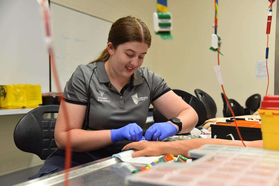 A paramedic student sits at a clinical table demonstrating how to insert a canula into a dummy arm. IV bags hang from above, and the student has their medical equipment within reach on the bench.