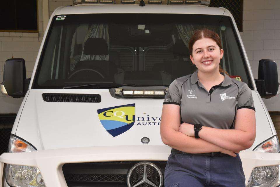 A paramedic student stands smiling with arms folded in a relaxed pose, at the helm of a CQU-branded training ambulance.