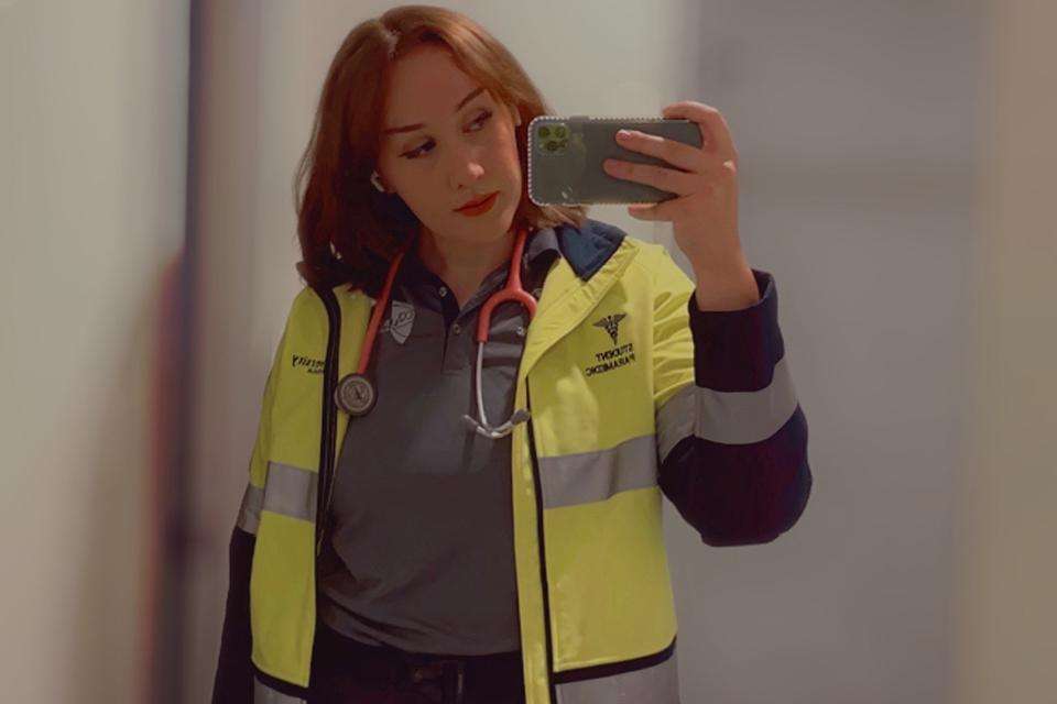 Student Paris Cox takes a selfie wearing her CQU paramedic science student uniform including hi-vis jacket and grey uniform shirt. She has shoulder length hair and also wears a stethoscope around her neck.