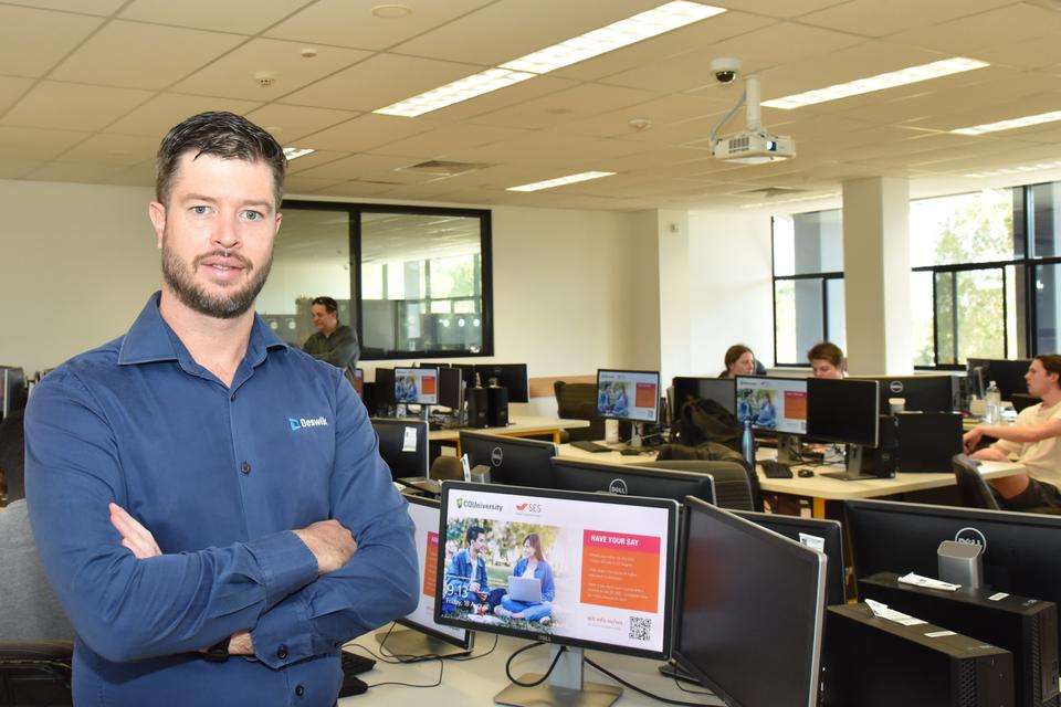 Patrick Doig stands facing the camera with arms folded in a blue shirt in a computer lab with students working behind him