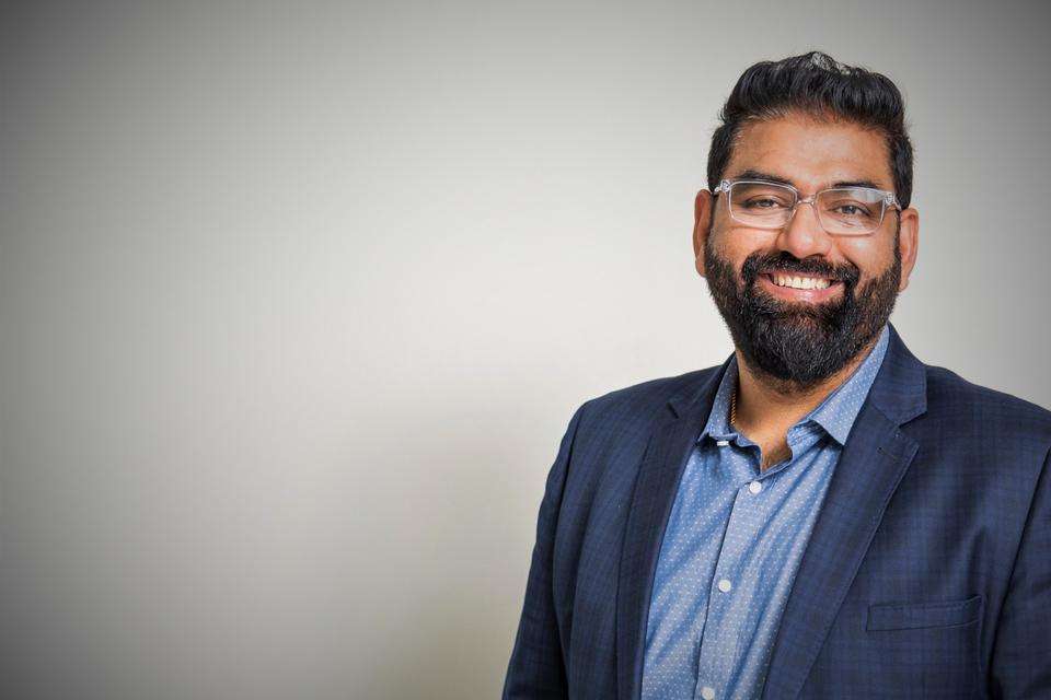 Man with short brown hair, olive skin, a beard wears clear glasses and stands to the right of screen wearing a light blue business shirt with navy blue jacket in front of a plain grey background