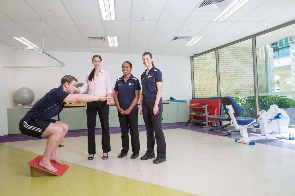 Three people look on while a person does a squat in a physiotherpy room