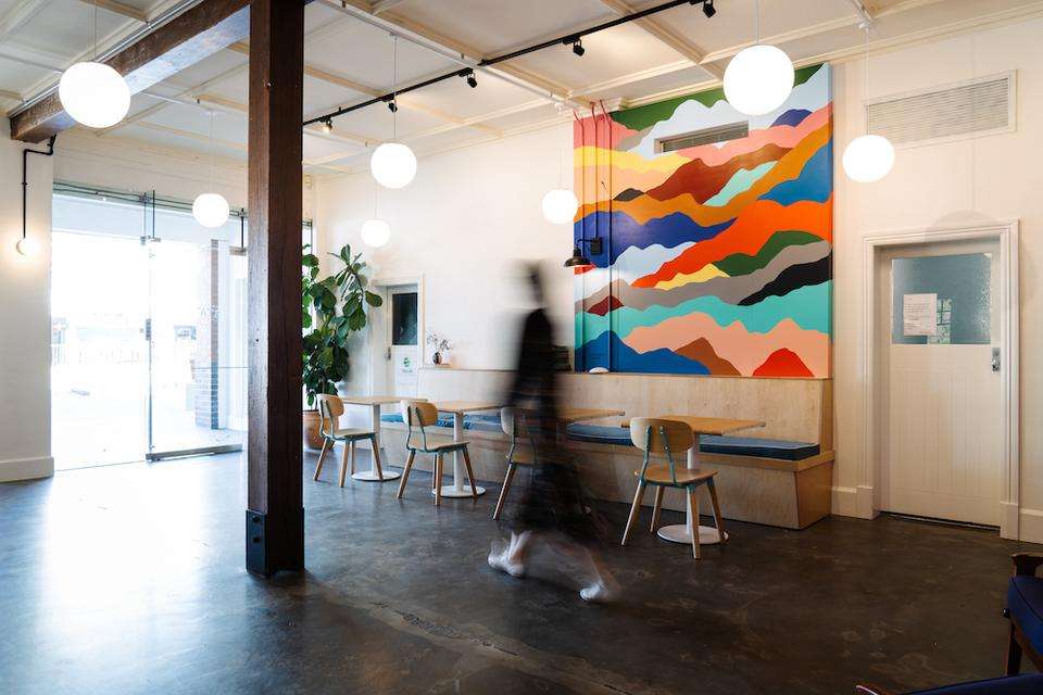 Inside of a cafe space with brightly coloured abstract artwork hanging on the wall, a person (blurred) walks through the cafe