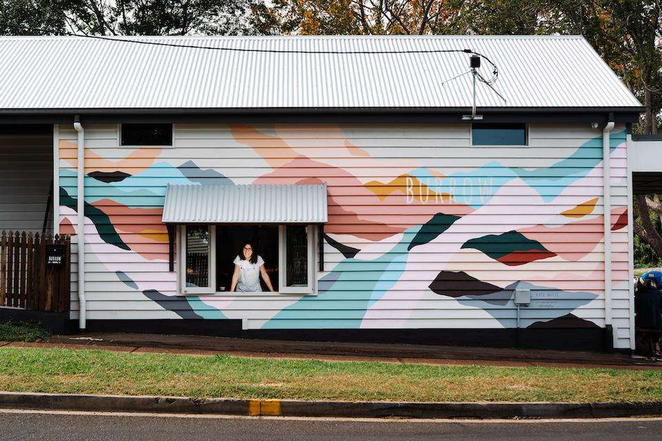 Photo of the facade of a country cabin painted brightly, with Visual artist Katie Whyte smiling from an open window