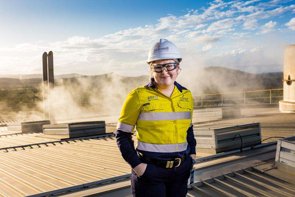A young woman in hard hat and hi-vis uniform with Stanwell logo stands on the roof of an industrial building with a mountain vista and chimney with steam in the background.