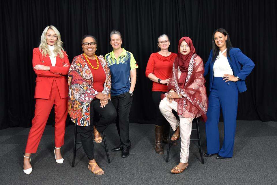 Six women dressed in bright, bold colours, all from various cultural backgrounds, in a studio, with a black curtain behind them, all looking directly at the camera and smiling