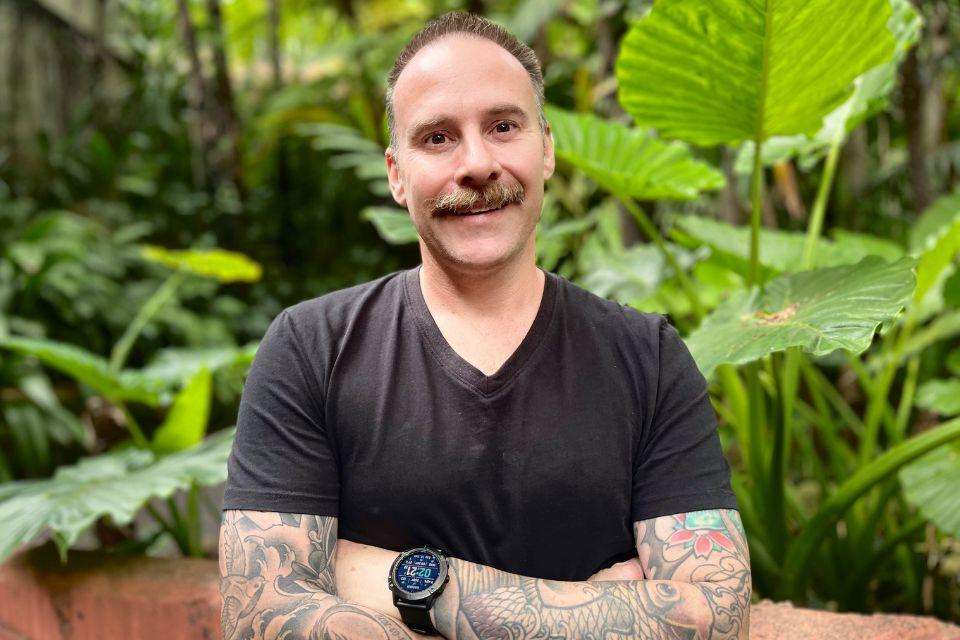 A man with a moustache, wearing a black tshirt, folding his tattooed forearms.