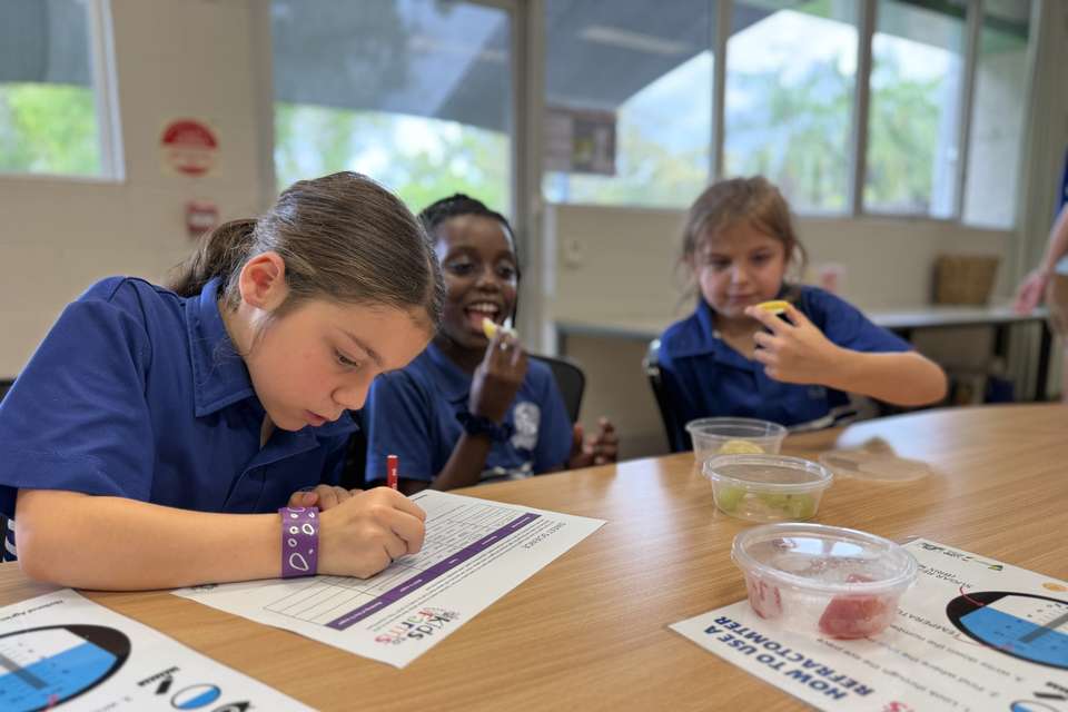 Three young children in school uniforms complete a classroom experiment looking at fruit. One child writes while two others eat a piece of fruit.