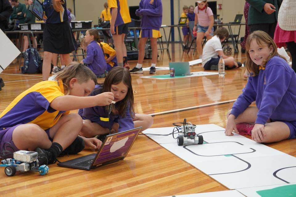 Primary students sit on the floor of a sports stadium with their mechanised robots, laptop and competition mat. Other students are pictured in various activity in the background.