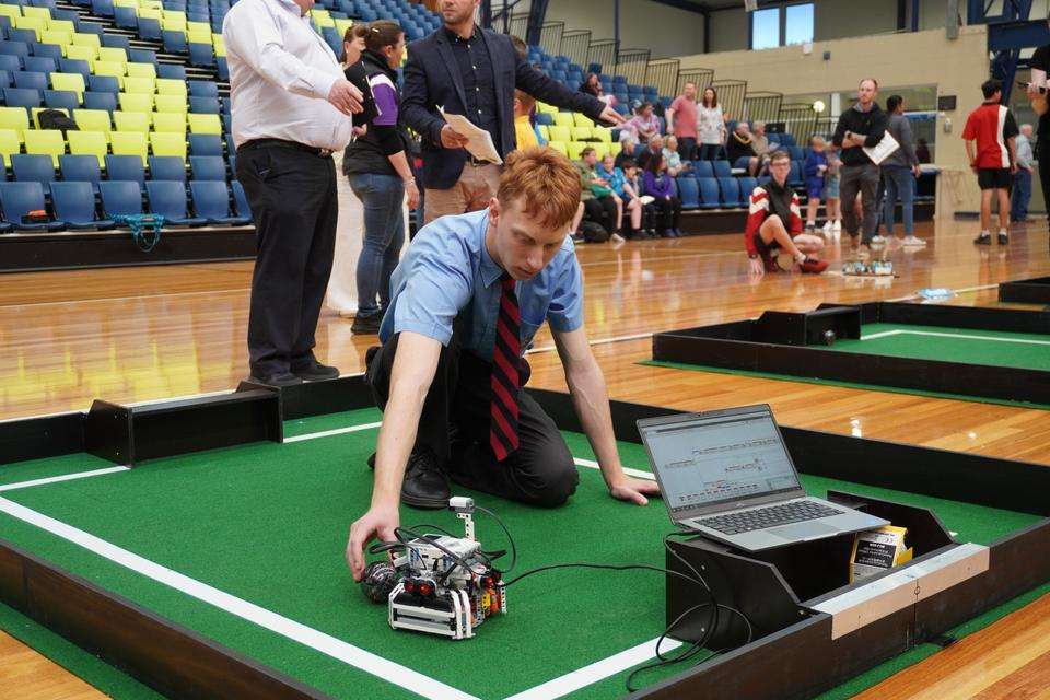 Competitor Daniel Spiryagin works with his robot on a replicated mini soccer field as he tests the sensors with a robotic ball and his laptop. Competition attendees and officials are in the background.